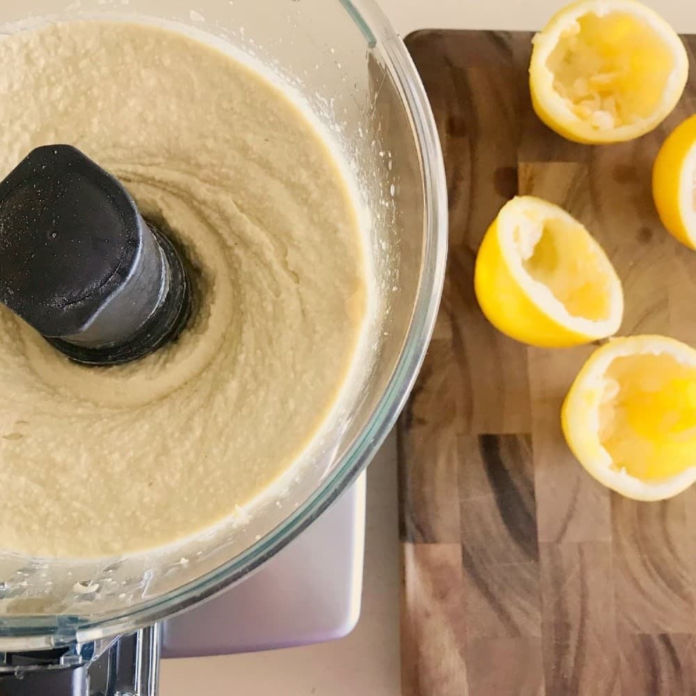 top view of a food processor bowl filled with creamy yellow garbanzo hummus, with wood cutting board and half lemons