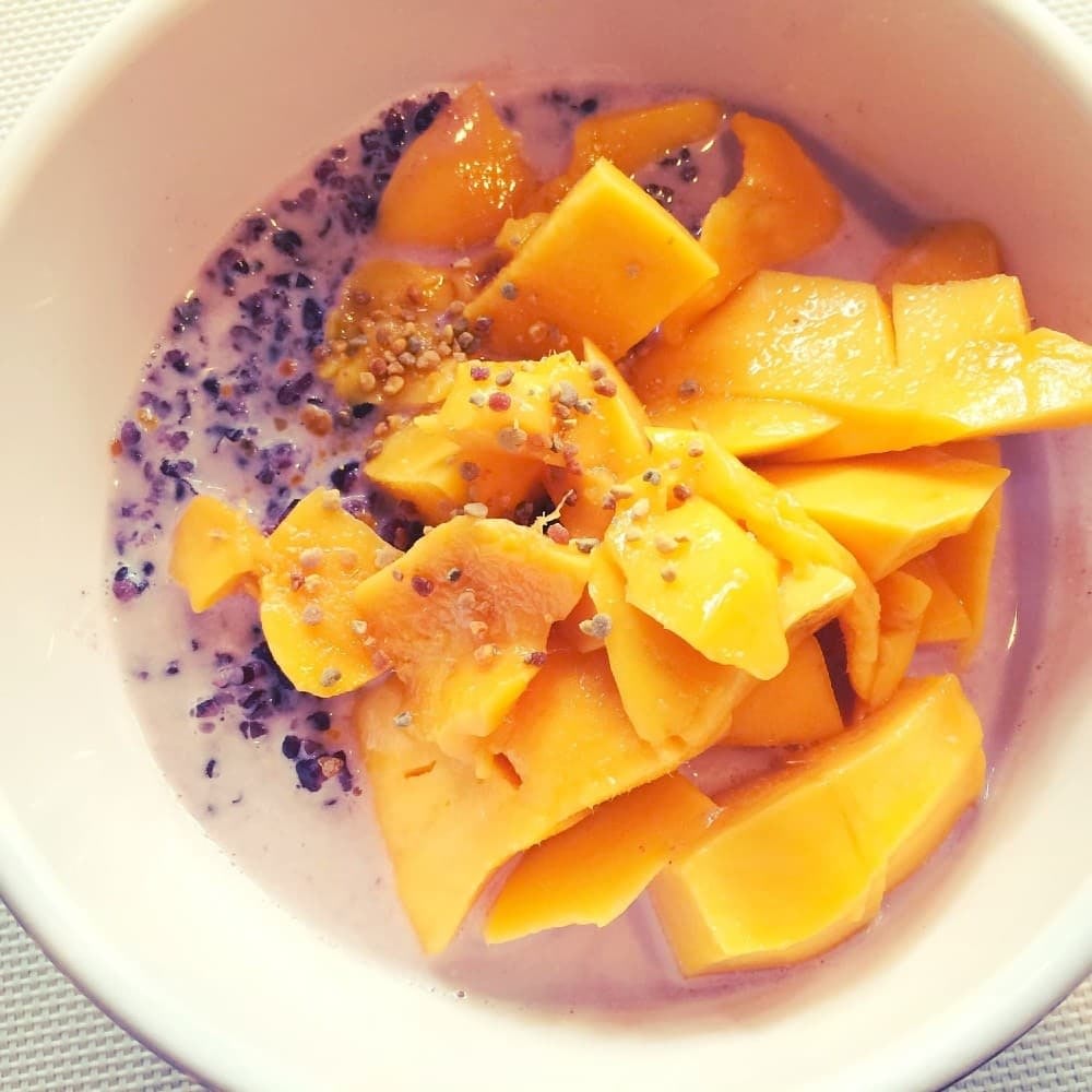 black rice porridge recipe, white ceramic bowl filled with cooked purple rice, creamy milk, and topped with fresh cut orange mango pieces
